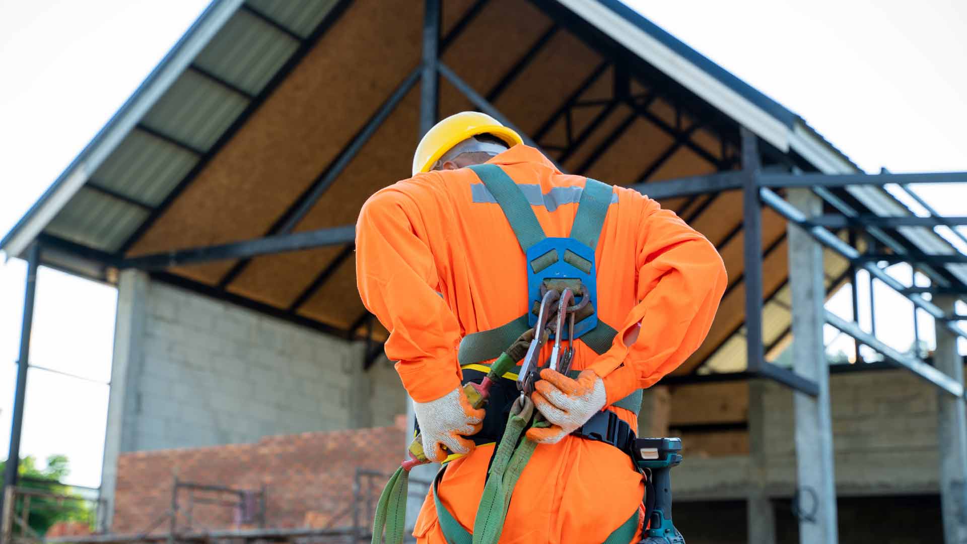 Vista posteriore di un operaio edile in un cantiere che indossa dispositivi di protezione individuale (DPI), tra cui una tuta arancione ad alta visibilità, un casco giallo e un'imbracatura di sicurezza con moschettoni, pronto per il lavoro in quota.