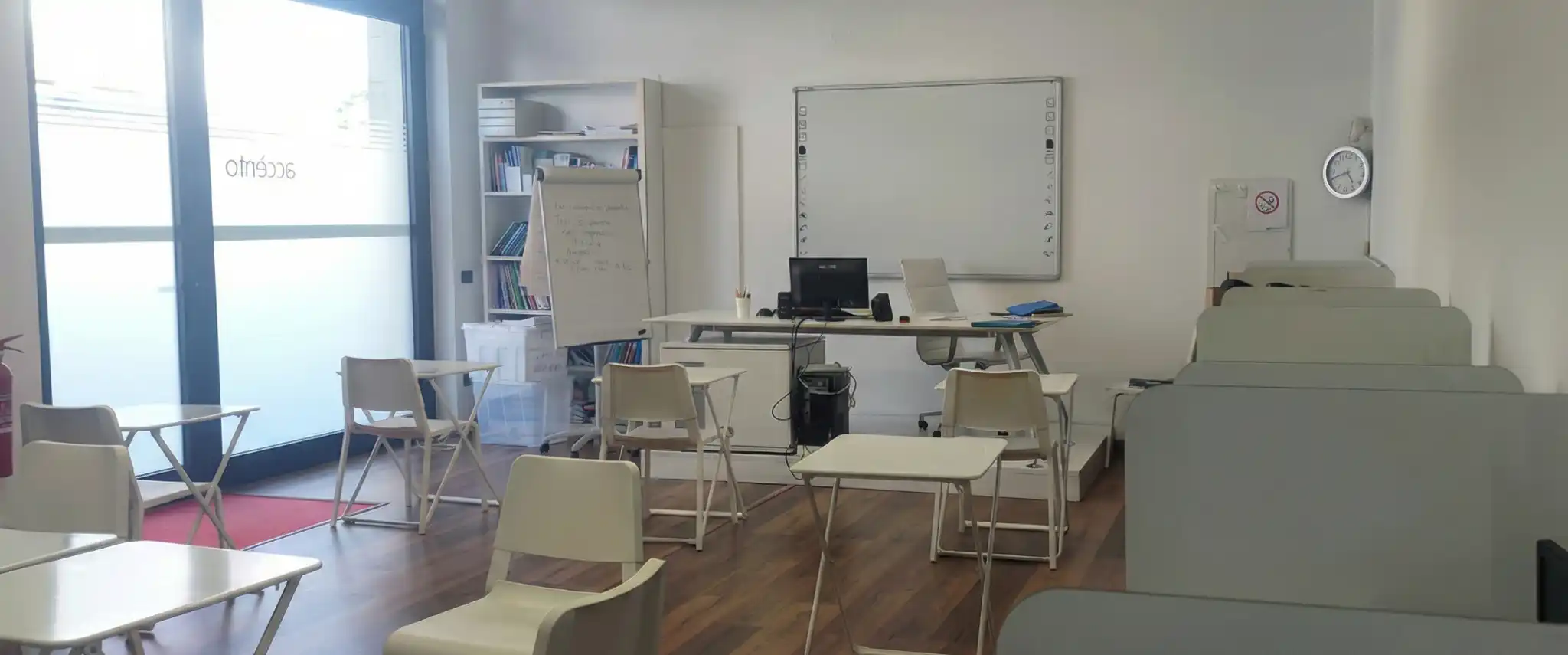 Panoramica di un'aula spaziosa con pavimento in parquet, arredata con file di sedie bianche con ribaltina, una cattedra con computer e una lavagna bianca sullo sfondo, pronta ad accogliere gli studenti di accento language academy.