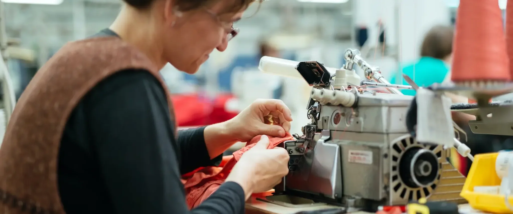 Vista laterale di una sarta industriale con occhiali che lavora con precisione su una macchina tagliacuci, assemblando un tessuto arancione in un laboratorio tessile.