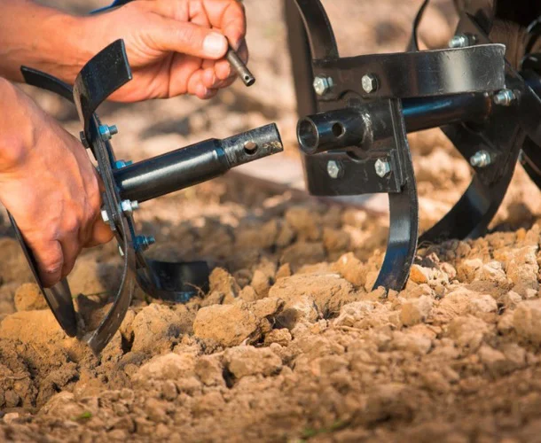 Dettaglio delle mani di un operatore che montano le lame di una motozappa inserendo un perno di fissaggio metallico, lavorando direttamente sul terreno agricolo.