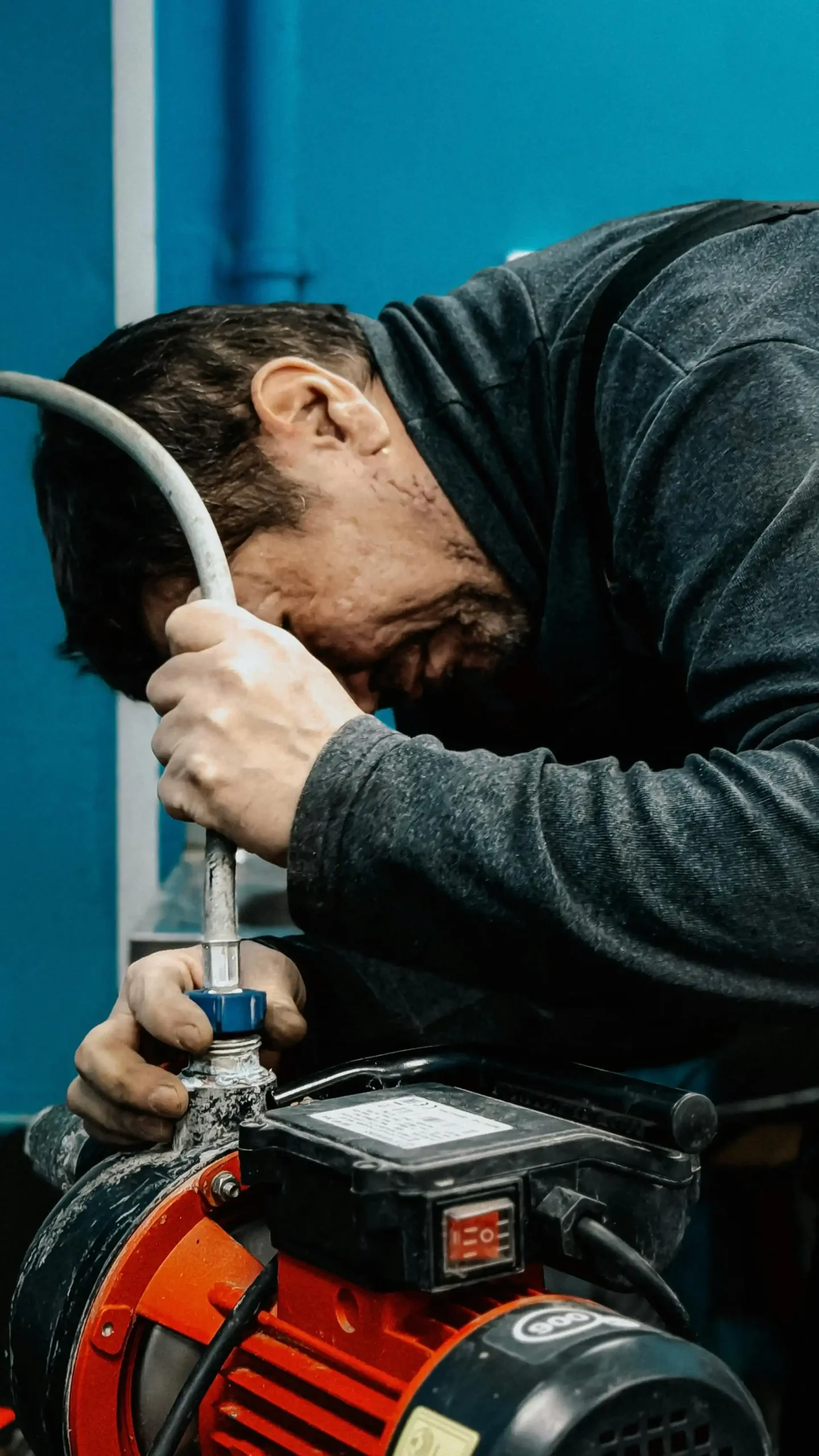 Un uomo di mezza età, con indosso una giacca da lavoro grigia scura, è inginocchiato e concentrato mentre stringe un tubo bianco all'attacco di una pompa dell'acqua arancione e nera. Si trova in un ambiente di lavoro, con una parete blu sullo sfondo.