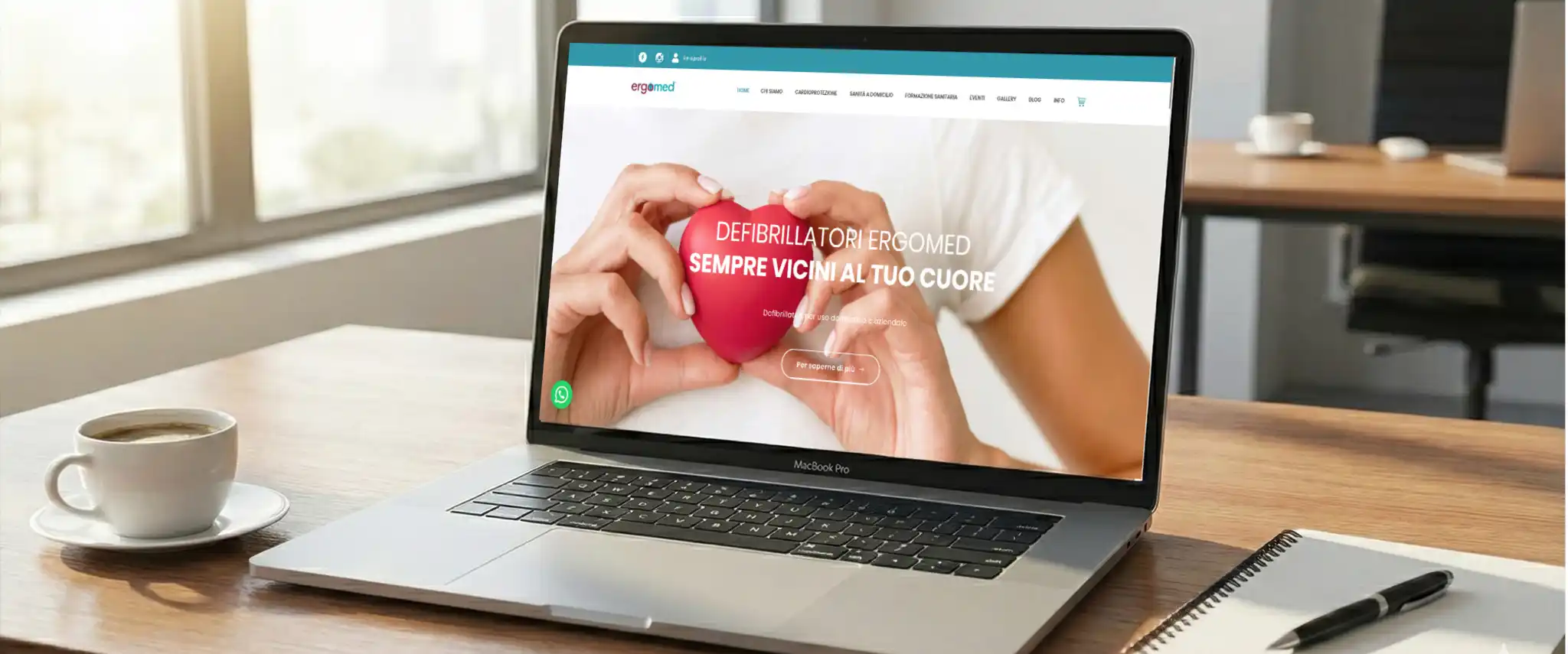 Un laptop aperto su una scrivania di legno visualizza la homepage del sito web ergomed.it, che presenta un'immagine di mani che tengono un cuore rosso con il testo "DEFIBRILLATORI ERGOMED SEMPRE VICINI AL TUO CUORE". A fianco del computer ci sono una tazza di caffè, un blocco note e una penna, con una luminosa finestra sullo sfondo.