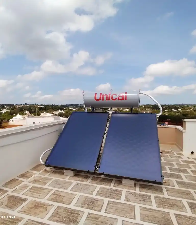 Un impianto solare termico marca Unical installato su una terrazza lastricata. Il sistema è composto da due collettori solari piani inclinati di colore blu scuro e un serbatoio di accumulo orizzontale grigio con logo rosso, posizionato sopra i pannelli. Sullo sfondo, un cielo azzurro con nuvole e un paesaggio rurale con vegetazione e abitazioni bianche.