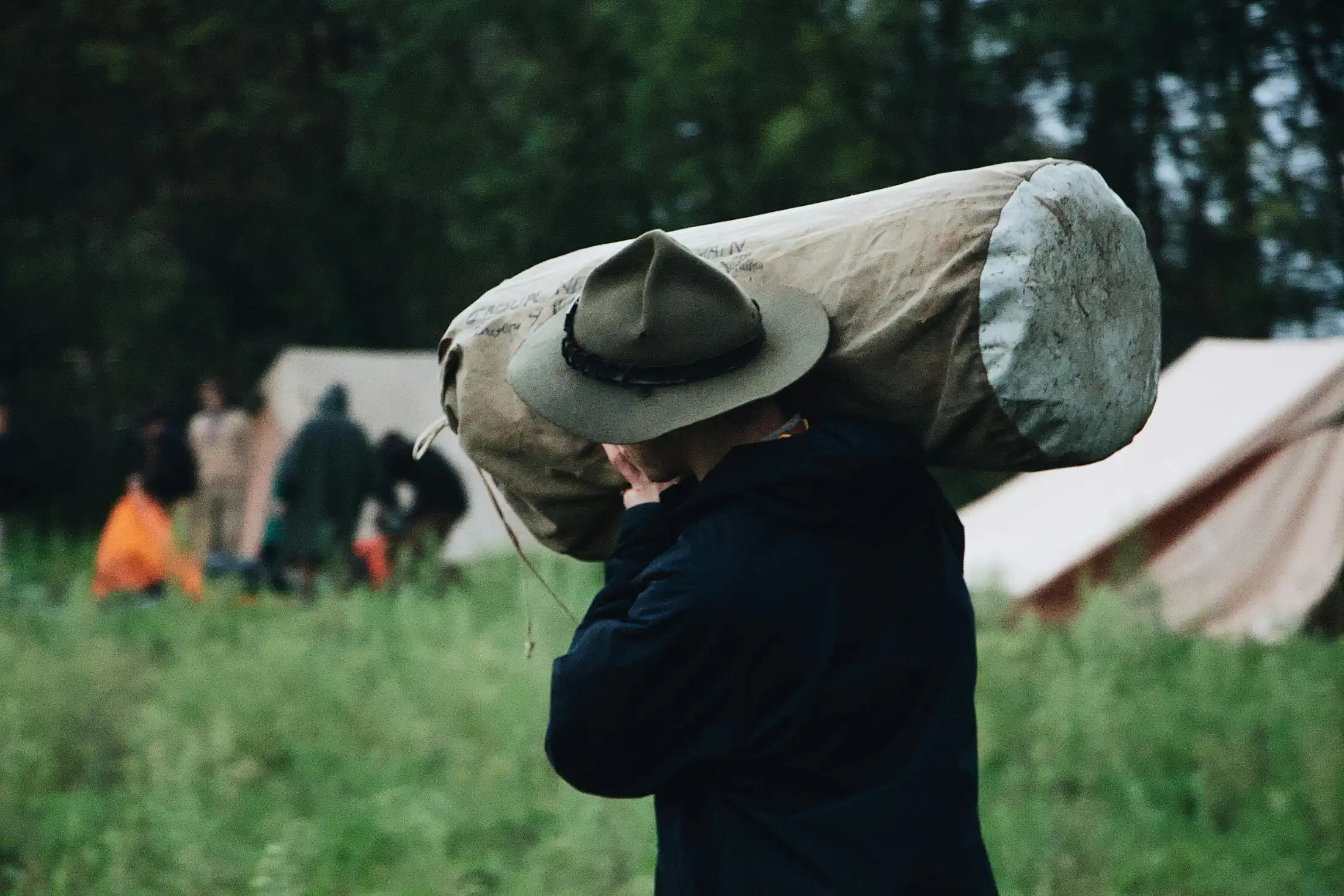 Vista di spalle di uno scout che indossa un cappello tradizionale e una giacca scura, mentre trasporta un grande e pesante sacco di tela contenente una tenda sulla spalla. Si trova in un campo erboso con altre tende visibili sullo sfondo sfocato, evocando la preparazione per un'avventura all'aria aperta.