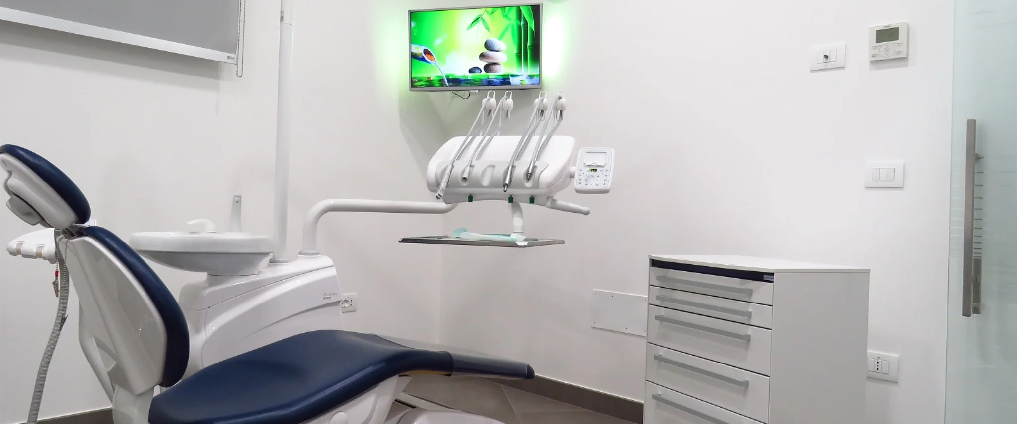 Panoramica di una sala visita dentistica luminosa e pulita, arredata con una poltrona operativa blu e bianca, cassettiera medica e un monitor a parete che proietta immagini rilassanti della natura per il comfort del paziente.
