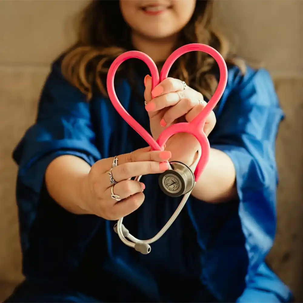 Le mani di una donna con unghie rosa e anelli d'argento tengono un fonendoscopio rosa, piegando i tubi per formare un cuore. La persona indossa un camice medico blu scuro.
